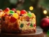 Fotografia de um pedaço de bolo de frutas, com uma crosta dourada e interior denso, adornado com frutas cristalizadas coloridas. O fundo desfocado apresenta luzes quentes.