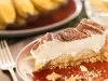 Em cima de uma mesa, fatia de banoffe com massa crocante, doce de leite e chantilly, decorada com canela em pó.