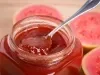 Fotografia de um pote de vidro decorativo com um doce de goiaba, na borda do pote tem uma colher de sobremesa apoiada com um pouco do doce. Ao redor do pote, que está sobre uma mesa de madeira clara, tem cinco metades de goiabas viradas pra cima.