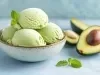 Imagem de um bowl claro com quatro bolas de sorvete de abacate decorado com uma folha de hortelã. Ao lado, abacates cortados ao meio.