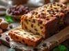 Em cima de uma mesa, tábua com bolo fofinho com frutas cristalizadas e uvas-passas pretas.