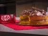 Fotografia em tons escuros de um bolo de cenoura com ganache de kitkat no recheio e na cobertura. As laterais do bolo estão sem a ganache, o bolo está apoiado em um pano vermelho sobre uma mesa cinza. Ao lado tem uma embalagem de kitkat.