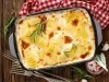 Em cima de uma mesa, refratário com batatas gratinadas com molho branco, decorado com alecrim.