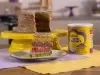 Fotografia em tons claros de um bolo inteiro sobre um apoio de bolo amarelo sobre dois panos, roxo e amarelo, em uma mesa de madeira. Ao lado do bolo, que está aberto com camadas coloridas, tem uma lata de NINHO Nutri Advance e uma fatia do bolo.