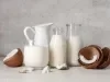 Fotografia de duas garrafas decorativas de vidro, uma jarra branca e um copo grande e largo. Todos esses recipientes têm bebida vegetal de coco. Ao lado tem cascas de coco pela metade, alguns pedaços e um pouco da polpa sobre uma mesa cinza clara.