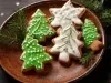 Em cima de uma mesa, prato redondo com biscoitos em forma de árvore de Natal e estrela, decorado com glacê branco e verde, e confeitos redondos brancos.