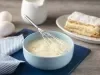 Fotografia em tons de azul em uma bancada de madeira, um pano azul listrado, uma tábua de madeira azul, um bowl azul claro com o creme belga dentro e um fouet. Ao lado, um prato com um pedaço de uma torta com o creme belga.