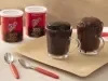Foto da receita de Bolo de chocolate na caneca servida em duas canecas de vidro sobre uma tábua de madeira com duas latas de nescau extra cacau e duas colheres de decoração