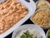 Fotografia de uam travessa branca retangular com o strogonoff de frango, ao redor tem três recipientes. Em cada um tem o champignon fatiado, batata palha e arroz branco.