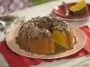 Imagem de um bolo de cenoura com cobertura de brigadeiro e raspas de chocolate sobre um prato rosa claro de bolinhas brancas. Ao fundo, um prato com uma fatia de bolo. u