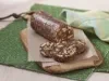 Fotografia em tons de verde em uma bancada de madeira com um pano verde florido, uma tábua de madeira com um papel bege e salaminho de chocolate.