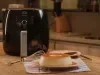 Imagem de um pudim de leite condensado Moça sobre um prato branco, ao lado de um fouet e uma air fryer preta, em uma bancada de cozinha.