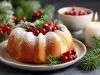 Fotografia de um bolo de Natal apresentado de forma festiva. O bolo, dourado e polvilhado com açúcar, é decorado com cranberries vermelhas e ramos de pinheiro. Ao fundo, uma vela acesa e mais cranberries complementam a atmosfera natalina.