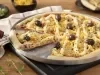 Fotografia em tons amarelo em uma bancada de madeira, uma tábua de alumínio redonda com a pizza de frango com catupiry em cima dela. Ao lado, potinhos com milho, azeitonas e um prato com uma fatia da pizza.