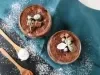 Fotografia vista de cima de dois copos de vidro decorativos sobre um peno de tom azul dobrado, com açúcar de confeiteiro polvilhado por cima do pano e da sobremesa. Dentro do copo tem a sobremesa, finalizada com ganache, uva e cacau em pó.