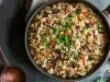 Em cima de uma mesa, frigideira com arroz soltinho uvas-passas pretas e brancas, decorado com folhas de cheiro-verde.