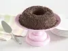 Foto de um brigadeirão de chocolate em cima de um prato de bolo rosa e com pratos brancos atrás