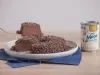 Foto da receita de Brigadeirão Moça, servido em um prato branco, decorado com muitos granulados de chocolate. O prato está entre uma lata de Leite Moça e uma espátula dourada, tudo sobre uma bancada de madeira