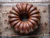 Fotografia de um bolo de chocolate em uma grade de resfriamento, com cor marrom escura e superfície brilhante. O bolo tem um padrão flutuante e está sobre uma superfície de madeira rústica.