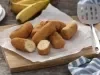 Fotografia em tons de amarelo e azul em uma bancada de madeira clara, um pano azul com listras brancas, uma tábua de madeira com várias bananas à milanesa em cima dela. Ao lado, uma concha de fritura e ao fundo um cacho de bananas.
