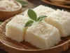 Fotografia de uma bandeja de bambu contendo quadrados de bolo de tapioca, cobertos com coco ralado, dispostos sobre uma superfície de madeira, com um fundo que destaca mais coco e folhas verdes.