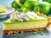 Fotografia em tons claros de uma fatia de torta de limão com gelatina de limão e um pouco de chantilly por cima. Ao fundo tem limões inteiros e um prato de vidro branco e raso com fatias de limão.