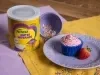 Fotografia de dois panos de mesa, roxo e amarelo, um sobre o outro em uma mesa de madeira. Sobre eles tem uma lata de NINHO Nutri Advance ao lado de um prato médio roxo com um cupcake e um morango inteiro, ao lado tem um potinho roxo com granulados.