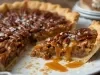Fotografia em tons claros de uma fatia de torta de doce de leite com nozes sobre um prato grande, raso e branco. A fatia está ao lado do restante da torta que está salpicada com flor de sal.