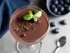 Fotografia de uma taça de vidro com a sobremesa de modo individual, finalizada com ganache, raspas de laranja, hortelã e mirtilos, para variar.