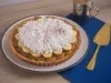 Receita de torta banoffee, servida num prato branco e decorada com chantilly e cacau em pó. A torta está ao lado de uma espátula dourada em uma bancada de madeira com um tecido na cor azul escura