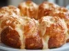 Fotografia de um bolo de pão de queijo, com pedaços douradas e brilhantes, cobertas por queijo derretido que escorre. O pão está em um prato branco, com um fundo suavemente desfocado.