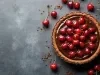 Fotografia vista de cima de uma torta inteira de chocolate e cerejas decorando a torta, que está dentro de uma forma de alumínio sobre uma mesa escura. Ao redor da torta tem cerejas inteiras.