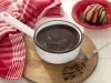 Fotografia em tons de vermelho de uma bancada, ao centro uma panela branca com uma calda de chocolate, em volta um pano vermelho e branco.