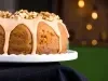 Bolo bundt elegantemente apresentado em um suporte branco. Com uma cobertura cremosa de cor caramelo e decorado com nozes picadas, o bolo exibe uma cor dourado-escura.