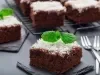 Fotografia de um quadrado de bolo de chocolate, coberto com coco ralado branco e decorado com uma folha de hortelã. O bolo está em uma tábua de ardósia escura, com mais pedaços ao fundo.