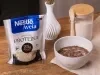 Receita de massa de brownie comestível servida em um pote branco sobre uma mesa de madeira decorada com um pote de aveia, um vaso e uma embalagem de nestlé aveia proteína