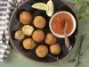 Fotografia em tons de verde em uma bancada de madeira verde, um pano listrado azul e cinza, um prato redondo preto com borda marrom e vários bolinhos de risoto em cima dele e um potinho branco com a geleia de pimenta para acompanhar.