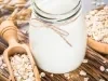 Fotografia em tons claros de um pote decorativo com bebida vegetal de coco sobre uma mini tábua de madeira escura retangular. Ao lado do pote tem aveia em flocos espalhadas e dentro de uma colher de madeira longa.