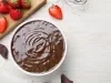 Fotografia em tons claros, visto de cima, de um recipiente redondo branco com a sobremesa, finalizada com ganache, sobre uma mesa de tom claro. Ao lado tem uma parte da tábua de madeira, retangular, com vários morangos, inteiros e cortados.
