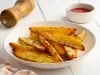 Fotografia em tons claros de um recipiente branco médio, fundo, sobre uma mesa de madeira branca. Dentro do recipiente tem batatas assadas e temperadas, cortadas em meia lua. Ao lado do recipiente tem temperos e ketchup em potinhos brancos.