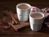 Foto dois copos brancos com chocolate quente dentro em cima de uma tábua de madeira com canela em pau e em pó decorando a mesa
