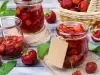 Potes de vidro com compota de morango, cercados por morangos frescos e cesta de frutas ao fundo.