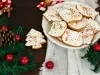 Em cima de uma mesa, prato redondo com biscoitos em forma de árvore e diversas formas, decorados com glacê branco e confeitos coloridos de bolinha.