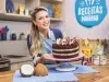 Imagem da confeiteira e influenciadora Beca Milano segurando a receita de bolo Naked de Prestígio atrás de uma bancada de cozinha, com pedaços de coco fresco e alguns produtos do lado direito