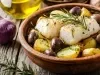 Em cima de uma mesa, refratário com pedaço de bacalhau, azeitonas pretas, batatas, decorado com alecrim.