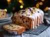 Fotografia de um bolo de frutas, com uma fatia cortada em destaque. O bolo é decorado com glacê branco e frutas secas coloridas. A cena é iluminada por uma vela, transmitindo um clima natalino.