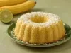 Fotografia de um bolo bundt de milho, dourado e coberto com coco, apresentado em um prato verde, com espigas de milho e uma lima ao fundo.