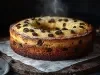 Fotografia de um bolo redondo dourado, exibido em uma tábua de madeira rústica. Com passas e uma leve camada de açúcar de confeiteiro, o bolo destaca-se em um fundo escuro.