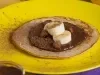 Fotografia de um prato amarelo com uma panqueca de banana com bananas por cima, creme de avelã e três aveãs inteiras. .