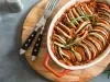 Fotografia em tons de vermelho com uma tabua de madeira ao centro. Em cima da tabua existe uma receita de ratatouille feita com berinjela, abobrinha, tomate e alecrim
